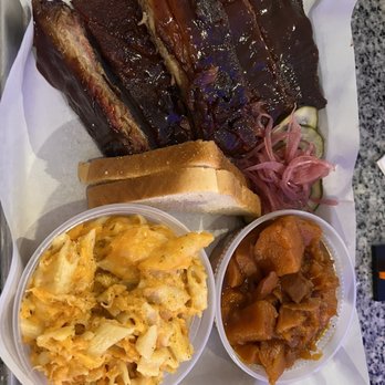 Backyard Barbeque - Pork Ribs  Smoked Mac & Cheese Smoked Candied Yams - A tray of barbecue ribs with a side of macaroni and cheese, baked beans, and pickles, served with slices of bread, at a restaurant. - Barbeque Near Me - Freeport, New York