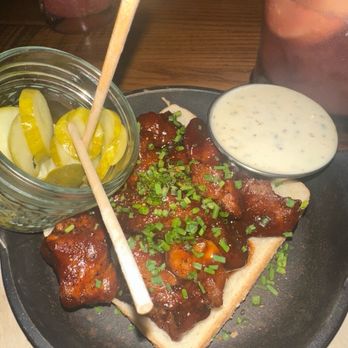 HangOverEasy - A black plate holds a serving of barbecue chicken topped with chopped green onions, accompanied by a small bowl of creamy white dipping sauce. There are pickles in a glass jar with a stick, and two glasses of a pink beverage in the background. The setting appears to be a restaurant with a wooden table surface. - Breakfast & Brunch Near Me - Cincinnati, Ohio