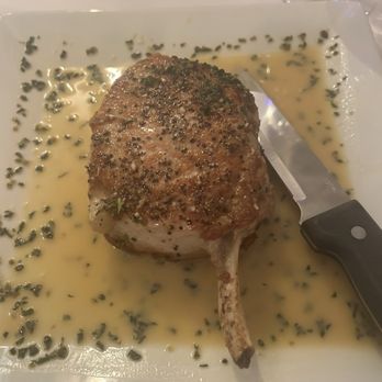 Pete's Taverna - The BEST pork chop EVER. - A pork chop seasoned with herbs and spices is served on a white plate with a sauce and chopped herbs, accompanied by a knife. - Cocktail Bars Near Me - Riverhead, New York