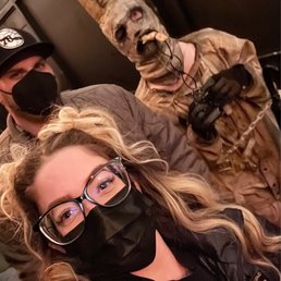 A woman with glasses and a black face mask, with blonde hair styled in loose waves, takes a selfie with two individuals in costumes. One person is dressed in a horror-themed costume with a skull mask, a long pointed hat, and a brown outfit, holding a camera. The background suggests an indoor setting, likely at Fright Haven haunted house attraction.
