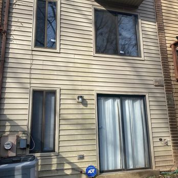 Montgomery Decks - Before. - The image shows the exterior wall of a beige residential building with horizontal siding. There are four windows: two small vertical windows on the left, one larger window on the upper right, and a sliding glass door with curtains on the lower right. An ADT security sign is placed near the ground, and utility meters are mounted on the wall. The ground has some leaves and a concrete step in front of the sliding door. - Decks & Railing Near Me - Frederick, Maryland
