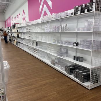 Daiso - household items - The image shows a retail store aisle with mostly empty white metal shelves. Various plastic storage containers, organizers, and boxes are placed on the shelves, mostly in clear and black colors. The store has a pink wall with white geometric arrow designs and the word 'Daiso' visible in the background. The floor is wood-patterned, and a person is seen walking in the distance. - Department Stores Near Me - Olathe, Kansas