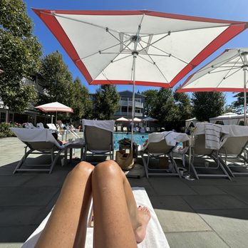 Harbor View Hotel - The pool has cabanas and plenty of lounge chairs. Access to full kitchen menu. - The image shows a person lying on a white towel on a lounge chair beside a swimming pool. Large umbrellas provide shade, and other lounge chairs with towels and people are visible around the pool area. The background includes trees, residential buildings, and a bright, sunny sky, indicating a warm, outdoor setting at a hotel or resort. - Hotels Near Me - Edgartown, Massachusetts