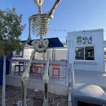 The Block - The Block.  Halloween themed skeleton. - A large skeleton decoration with an outstretched arm stands outdoors on a patch of mulch, near a building with a sign that says 'Tiki Tiki' and features a pineapple graphic. The skeleton is made of plastic or similar material, with detailed bones and a skull. It is positioned next to a small tree and is surrounded by some discarded packaging. The scene is set under a clear blue sky, with string lights hanging above the building, indicating a festive or themed outdoor area. - Shopping Centers Near Me - Rio Rancho, New Mexico