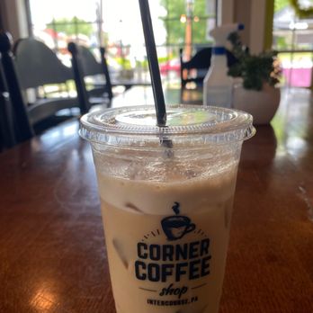 Corner Coffee Shop - A plastic cup containing iced coffee with ice cubes and a black straw is positioned on a wooden table inside a coffee shop. The cup has the logo and name 'Corner Coffee shop' with the website 'intercourse.pa'. The background shows a window with natural light, chairs, and a spray bottle on the table, indicating an indoor setting. - Coffee & Tea Near Me - Intercourse, Pennsylvania