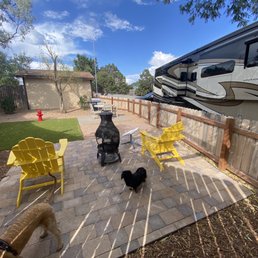 Private dog park / living area - The image shows a backyard patio with a stone-paved surface, containing yellow Adirondack chairs, a black fire pit, and two dogs, one black and one tan. The patio is enclosed by a wooden fence, with a RV parked adjacent to it. In the background, there is a small grassy area with a fire hydrant, a shed, and some trees under a partly cloudy sky. The scene appears to be part of a private outdoor space at a campground or RV park.