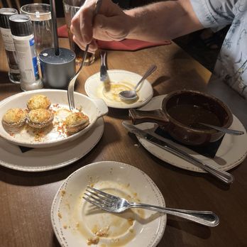 Seared - Stuffed mushrooms - A wooden dining table holds several dishes and utensils, including a partially eaten plate of stuffed mushrooms, an empty plate with a fork, a bowl of dark soup with a spoon, and a small plate with remnants of sauce. Condiments and salt and pepper shakers are also present. An arm with a short-sleeved shirt is reaching for food with a fork. The setting appears to be a restaurant with dim lighting and a casual atmosphere. - Steakhouses Near Me - Toledo, Ohio