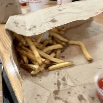 Five Guys - The "small" fry!!!! - A portion of French fries is served on a paper-lined tray, resting on a wooden table. The fries are golden-brown and slightly irregular in shape. In the background, there are cups with red and white branding, and other restaurant items, indicating a casual dining setting. - Burgers Near Me - Phoenix, Arizona
