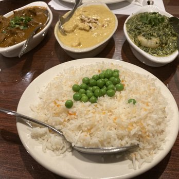 Namaste - Namaste.  Fish Korma, Shrimp Saag, Lamb Curry, and Jasmine rice and peas. - A large white plate of cooked white rice topped with green peas is placed on a wooden table. Surrounding the plate are three bowls containing Indian cuisine: one with a brown curry, another with a yellow curry, and a third with a green leafy vegetable dish. Each bowl has a spoon for serving. The setting suggests a shared meal at an Indian restaurant. - Indian Near Me - Rio Rancho, New Mexico