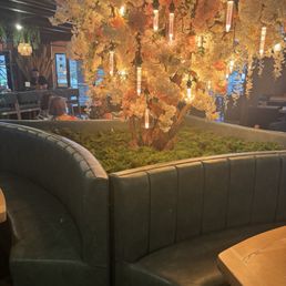 ambiance seating - A decorative artificial tree with white and pink flowers and hanging Edison-style filament bulbs is situated in a restaurant's seating area. The tree is planted in a circular booth with dark green leather upholstery, surrounded by tables and patrons. The background shows large windows and additional seating, creating a cozy ambiance.