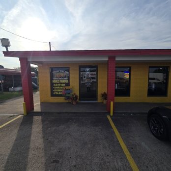 Fish Tales - The image shows the storefront of Fish Tales in Corrigan, TX, with a yellow brick facade and red-painted columns. The entrance has a glass door with signage, and a window displays a menu listing seafood items such as catfish, shrimp, gumbo, and po' boys. The parking lot in front has marked spaces, including one occupied by a black sports car. The sky above is partly cloudy, and there are additional structures and vehicles visible in the background. - Comfort Food Near Me - Corrigan, Texas