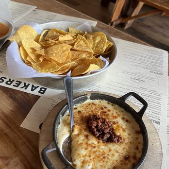Bakersfield Mass Ave - Chips and Chorizo Queso - A baked cheese dish topped with ground beef is served in a small cast iron skillet, accompanied by a bowl of tortilla chips on a restaurant table. - Bars Near Me - Indianapolis, Indiana