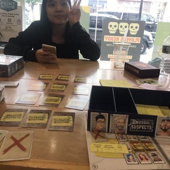 Squarrel Cafe - Yelp Celeb playing games with me - A woman with long dark hair sits at a table in a cafe, holding a phone in one hand and making a peace sign with the other. The table has game cards and a box for 'Unusual Suspects.' - Coffee & Tea Near Me - Brooklyn, New York