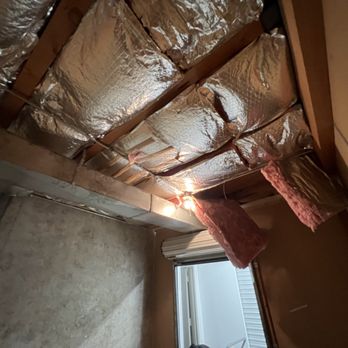 Public Storage - This is climate controlled and not considered a fire hazard by Public Storage. That's not what gov't experts believe. - Ceiling with exposed wooden beams and joists, featuring reflective silver insulation material and pink fiberglass insulation batts hanging down. A small window with blinds is visible below the ceiling, and a light bulb provides illumination. The space appears to be under construction or renovation. - Self Storage Near Me - Phoenix, Arizona