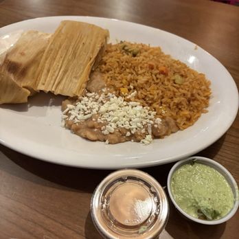 Quesa Tacos - Tamales - A white oval plate contains two tamales wrapped in corn husks, Mexican rice with vegetables, refried beans topped with crumbled cheese, and a side of green salsa. The plate is placed on a wooden table in a restaurant setting, with chairs and a pink chair back visible in the background. Two small containers of salsa and sauce are positioned in front of the plate. - Mexican Near Me - Carmel, Indiana