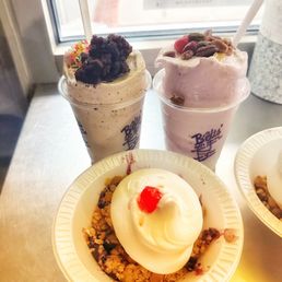 Flurries - Three soft serve ice cream desserts are shown on a metal counter near a window. The first is a vanilla-flavored treat topped with a blueberry crumble and colorful sprinkles. The second is a pink berry-flavored flurry with nuts and berries, served in a clear cup with a straw. The third is a vanilla crumb dessert with a dollop of whipped cream and a cherry on top, served in a small paper bowl. The background includes a window with a sign and a container of sprinkles.
