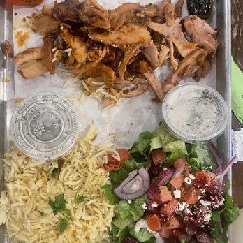 Meso Grill - Chicken plate without the bread - The image shows a metal tray containing a Middle Eastern meal. It includes shredded cooked chicken, a portion of seasoned rice, a fresh salad with lettuce, tomatoes, onions, and black olives, and roasted peppers. Two small clear plastic containers with lids are also on the tray, likely holding sauces or dressings. The tray is placed on a wooden table, with some papers partially visible underneath. - Mediterranean Near Me - Albuquerque, New Mexico