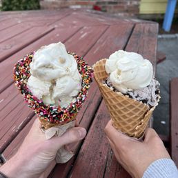 Chocolate Dipped Waffle Cone - Two ice cream cones are held by different individuals over a weathered wooden table outdoors. The left cone has a scoop of vanilla ice cream with a colorful sprinkle-coated rim, while the right cone contains a scoop of vanilla ice cream in a waffle cone. The background includes a brick wall, a yellow building, and a blue mailbox, indicating an outdoor setting.