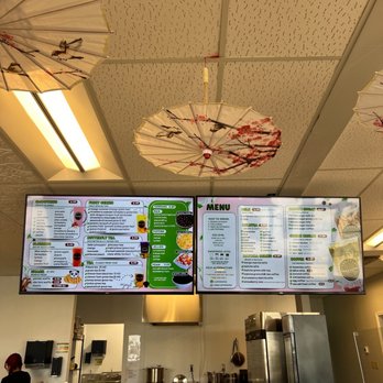 Teazentea - Menu - Ceiling features decorative paper umbrellas with cherry blossom patterns hanging above digital menu screens at a bubble tea shop. The menu screens display various drink options, including teas, smoothies, and toppings. The shop interior includes kitchen equipment, a refrigerator, and a staff member visible in the background. - Bubble Tea Near Me - Portage, Indiana