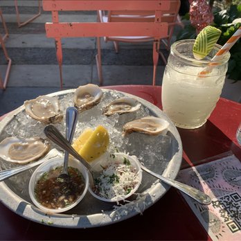 OTB - Oysters and frozen margarita - Oysters are arranged on a round metal tray filled with ice, with lemon wedges placed in the center. Two small bowls contain sauce and shredded garnish. A cocktail in a glass jar with a striped straw and a lime wedge is also on the table. The setting is outdoors with pink chairs and a patio background, suggesting a casual dining environment. - New American Near Me - Brooklyn, New York