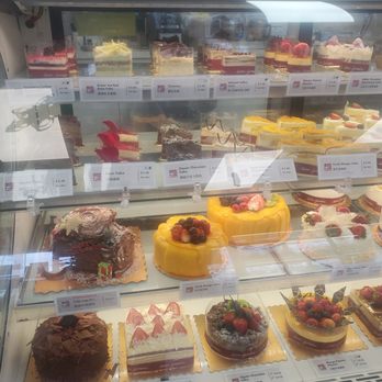 Adonis - A bakery display case contains multiple tiers of cakes and desserts, including fruit-topped cakes, chocolate cakes, and layered pastries. Labels with names and prices are visible, indicating a variety of flavors such as raspberry lychee, green tea red bean, tiramisu, and mango mousse. The desserts are arranged neatly, with some decorated with fruit, chocolate, or whipped cream. The case is made of glass, allowing a clear view of the items inside. - Grocery Near Me - Toronto, Ontario