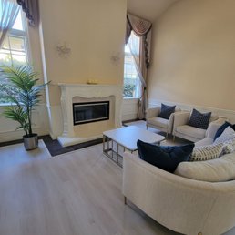 Formal livingroom spotless and windows wiped down very well. - A living room features a white fireplace, two large windows with curtains, and beige sofas with decorative pillows, creating a cozy space.