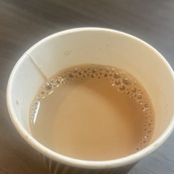 Tabla - A paper cup filled with chai tea, showing a light brown color with bubbles on the surface, sits on a wooden table. The cup appears to be made of corrugated paper, and the background includes a blurred object, possibly a tray or container. The image captures a close-up view of the beverage in a casual setting. - Indian Near Me - Phoenix, Arizona