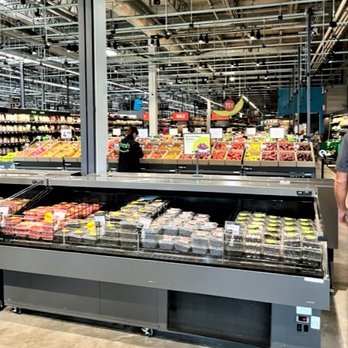 Amazon Fresh - Hot bar, salad bar, & more - A grocery store interior features a refrigerated display case filled with packaged food products, with shelves of fresh produce visible in the background. - Grocery Near Me - Oceanside, New York