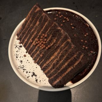 6Smith - Chocolate cake - A slice of layered chocolate cake with multiple thin layers of chocolate sponge and frosting, topped with small chocolate sprinkles. The cake is served in a white bowl, with some crumbs and chocolate dust on the side. The background includes a dark table surface and a white plate with a fork. - Seafood Near Me - Wayzata, Minnesota