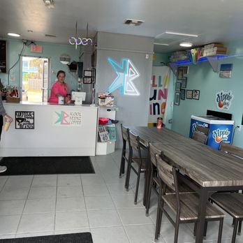 Kool Beenz Coffee - Place to sit inside - The image shows the interior of Kool Beenz Coffee, a family-owned cafe, featuring a wooden table with multiple chairs, a counter with a staff member in a pink shirt, neon signs including an 'open' sign and a stylized logo, and wall decorations such as framed pictures and colorful artwork. The space has tiled flooring, ceiling vents, and shelves with supplies, creating a casual and welcoming atmosphere. - Coffee & Tea Near Me - Flagler Beach, Florida