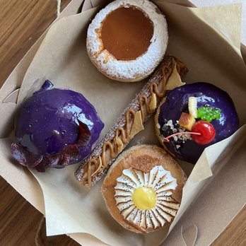 Kora - leche flan ni lola (upper left) , ube (bottom left) kalamansi amapola (bottom right), halo-halo (upper right), and churron (middle) - A cardboard box contains five Filipino-inspired desserts and pastries. There is a caramel-topped pastry dusted with powdered sugar, two purple glazed treats decorated with toppings including a cherry and candy, a long pastry with caramel drizzle, and a tart with a yellow center and decorative icing. The desserts are arranged on parchment paper inside the box, which is placed on a wooden surface. - Filipino Near Me - Sunnyside, New York