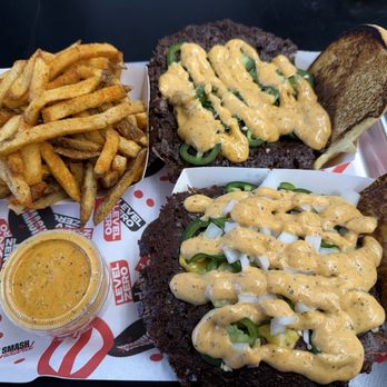 Level Zero - The image shows a tray with two smash burgers topped with jalapeños, onions, and a creamy sauce, each with toasted buns on the side. A portion of French fries and a small container of dipping sauce are also present on the tray. The tray is lined with branded paper from a burger restaurant. - Halal Near Me - Dearborn, Michigan