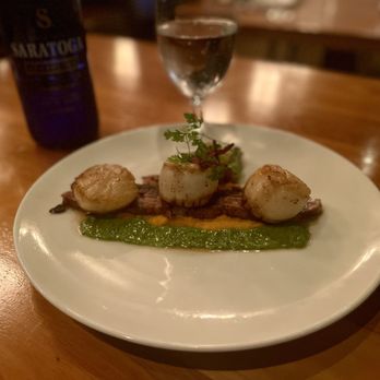 Scratch - Seared Scallops - Seared scallops with a green sauce on a white plate, garnished with herbs, in a restaurant setting with a wooden table. - New American Near Me - Mountain View, California