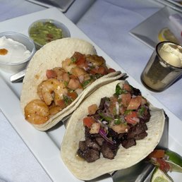 Tacos - Two tacos filled with shrimp and grilled beef, topped with pico de gallo, served on a white platter with lime wedges, guacamole, sour cream, and a beverage.