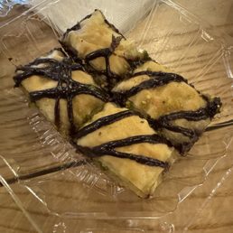 Chocolate buklawa - Four pieces of baklava with a flaky, layered pastry and chopped pistachios are arranged in a clear plastic container. Each piece is decorated with a drizzle of dark chocolate on top. The container is placed on a wooden surface, with a paper napkin and a stack of plastic water bottles visible in the background.