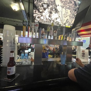 Kelly's Bleachers - What's on tap - A bar interior featuring multiple beer taps, a television displaying a football game, and patrons seated at the bar. - Sports Bars Near Me - Milwaukee, Wisconsin