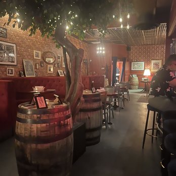 CODE RED - A dimly lit bar interior features a large tree growing through two wooden barrels used as tables. The brick wall behind is decorated with framed photos and artwork, creating a vintage ambiance. Warm lighting from hanging bulbs and a chandelier adds to the cozy atmosphere, with a person seated at the bar on the right side. - Cocktail Bars Near Me - Washington, DC, District of Columbia