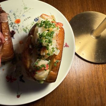 6 NYC - Surf and Turf Slider (Lobster Roll) - A lobster roll with chopped green onions on top is presented on a white plate with a floral pattern. The roll contains lobster meat and is served on a wooden table. A metal stand is visible to the right of the plate, suggesting a restaurant setting. - New American Near Me - Brooklyn, New York