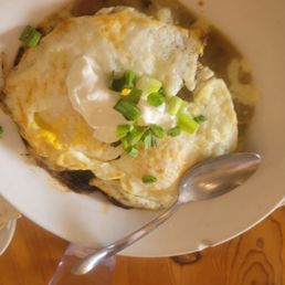 Huevos Rancheros - A bowl contains cooked eggs topped with chopped green onions and a dollop of sour cream. The eggs appear to be fried or scrambled, with some browning visible. The bowl is placed on a wooden table, and a metal spoon rests inside it. Part of another dish, possibly tortillas, is visible in the corner of the image.