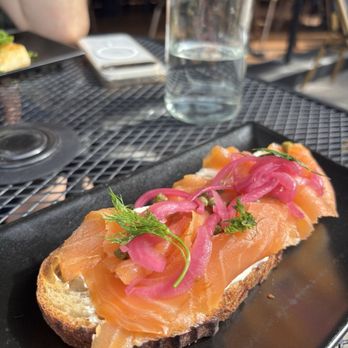 The Corner Store - Smokes Salmon toast - A close-up of smoked salmon toast garnished with pickled onions and herbs, served on a black plate at a restaurant table. - Coffee & Tea Near Me - Crown Heights, New York