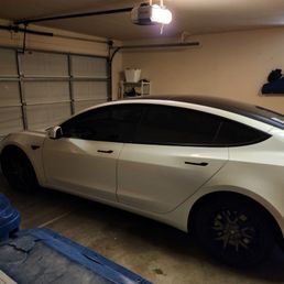A white Tesla Model S with tinted windows is parked in a garage with a closed sectional door, shelves, and storage items visible.