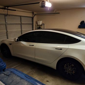 On Site Window Tinting - A white Tesla Model S with tinted windows is parked in a garage with a closed sectional door, shelves, and storage items visible. - Car Window Tinting Near Me