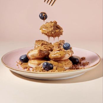 Chocolate Bash - A dessert presentation shows a stack of éclairs topped with blueberries and drizzled with caramel sauce on a pink-rimmed plate. A gold-colored fork is positioned above, dropping a blueberry onto the éclairs. The background is a plain, light-colored surface, emphasizing the dessert's details and textures. - Desserts Near Me - Manteca, California
