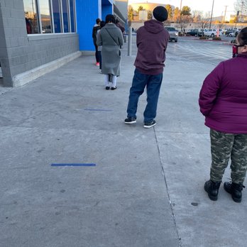Walmart Supercenter - Several individuals are waiting in line outside a retail store, maintaining social distance, in a parking lot with cars and trees in the background. - Grocery Near Me - Albuquerque, New Mexico