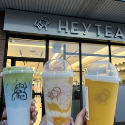 Three individuals display different beverages with transparent lids and logos, standing outside the HEYTEA store in Toronto, ON.