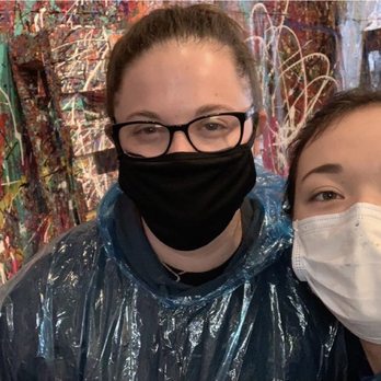 The Splatter Studio - Two women wearing masks, one with glasses and a black mask and the other with a white mask, are taking a selfie in front of a vibrant, abstract splatter painting. They are dressed in protective plastic smocks, indicating they are participating in a painting activity at The Splatter Studio in Atlanta, GA. The background features a wall covered with multicolored paint splatters and several chairs and art supplies. - Team Building Activities Near Me - Atlanta, Georgia
