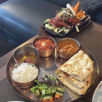 Curry Kitchen - Curry Dinner Special Combo for 1 - The image shows a metal tray containing naan bread, steamed white rice, and three small bowls of different Indian curries, including a tomato-based, a creamy brown, and a red sauce. A side salad with mixed greens, cucumber, and carrots is also present. In the background, a black plate displays grilled meat topped with vegetables and garnishes. The setting appears to be a restaurant table with a dark placemat. - Indian Near Me - Chicago, Illinois