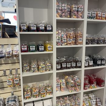Old Crows Antiques - White shelving units contain multiple rows of clear plastic bags filled with different types of candies, including gummies, chocolates, and sour candies, arranged neatly by color and variety. The store interior features natural lighting and other product displays nearby, indicating a retail environment focused on confectionery products. - Antiques Near Me - Golden, Colorado