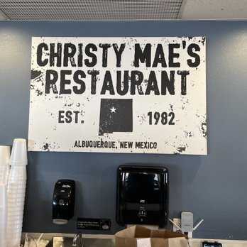 Christy Mae's Restaurant - Old sign - A rectangular sign with a distressed, vintage appearance displays the name 'Christy Mae's Restaurant' along with the establishment year, 1982, and the location, Albuquerque, New Mexico. The sign features bold black lettering and a black silhouette of the state of New Mexico with a star. Below the sign, there are disposable cups stacked on the counter, a black paper towel dispenser, and a small sign indicating employee handwashing requirements. The wall behind the sign is painted blue, and the ceiling has a perforated metal vent. - Sandwiches Near Me - Albuquerque, New Mexico