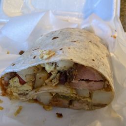 A breakfast burrito with visible potatoes, scrambled eggs, ham, and cheese, partially eaten and placed in a foam takeout container.